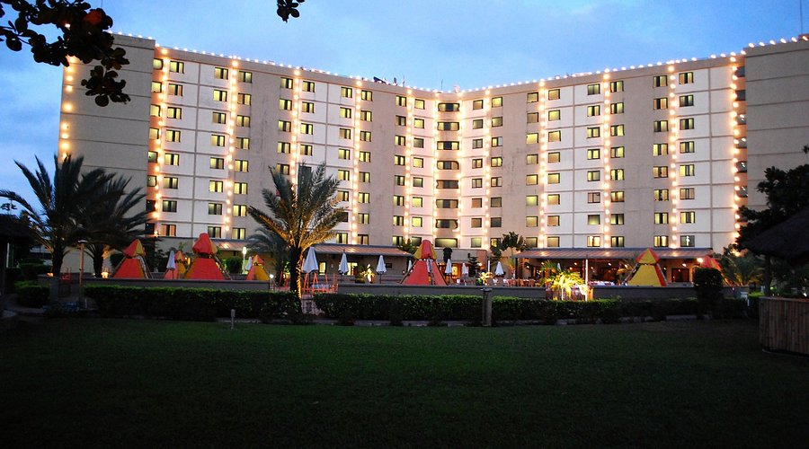 The Festival hotel curved white building illuminated by vertical string lights, with poolside cabanas and palm trees in foreground