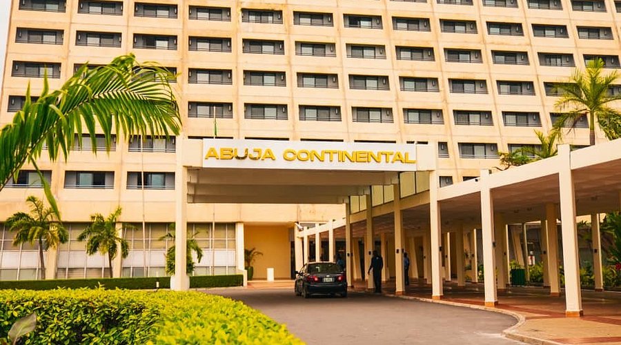 Abuja Continental Hotel entrance with covered driveway, palm trees, and “ABUJA CONTINENTAL” sign overhead